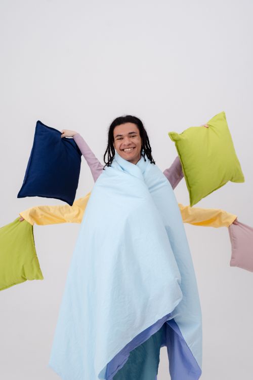 Free Smiling man wrapped in pastel blankets, holding vibrant pillows, on a white background. Stock Photo