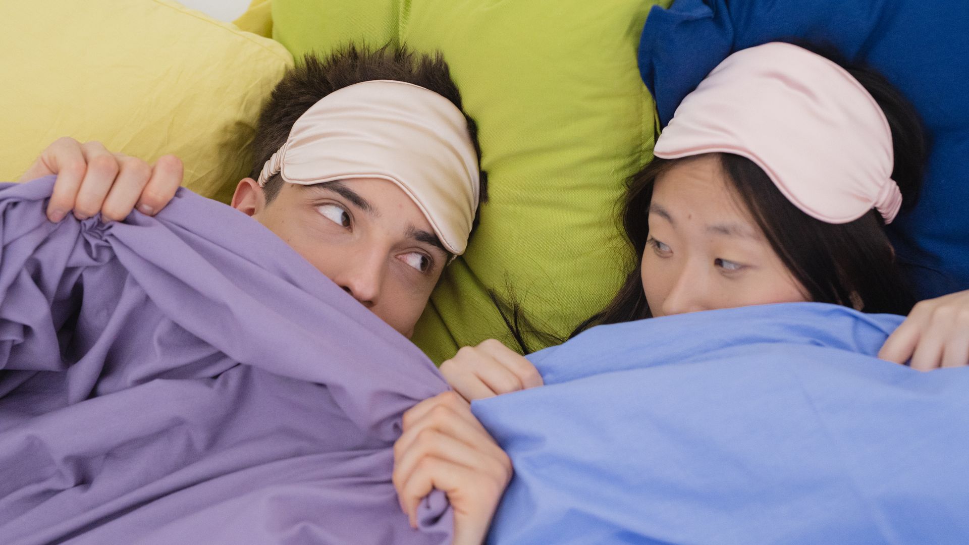 Modafinil Insomnia: Understanding the Link An interracial couple wearing sleep masks lying down on colorful pillows, portraying relaxation and rest — modafinil insomnia