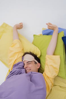 A relaxed man stretching in bed, wearing a sleep mask, under colorful sheets.