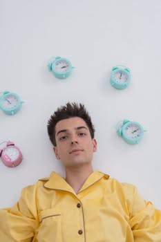 Portrait of a young man in yellow pajamas surrounded by colorful alarm clocks on a white background.