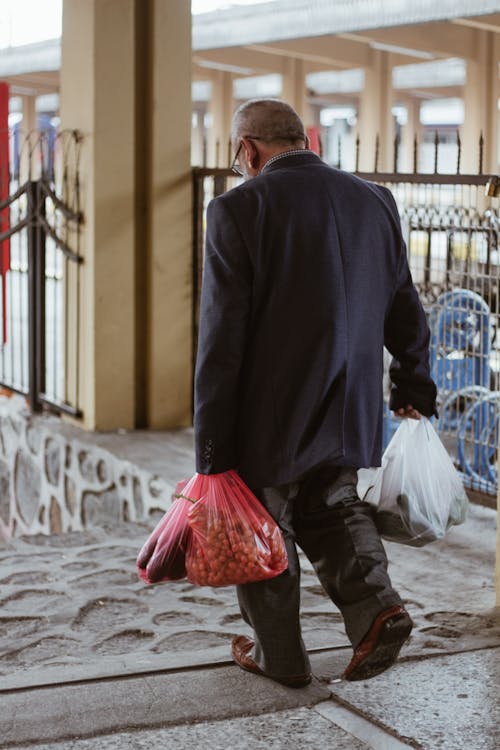 Back view of anonymous man with fresh fruits and vegetables in plastic bags strolling on walkway in town