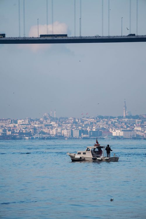 Free A motorboat cruises under a bridge in İstanbul with a scenic city skyline in the background, perfect for travel imagery. Stock Photo