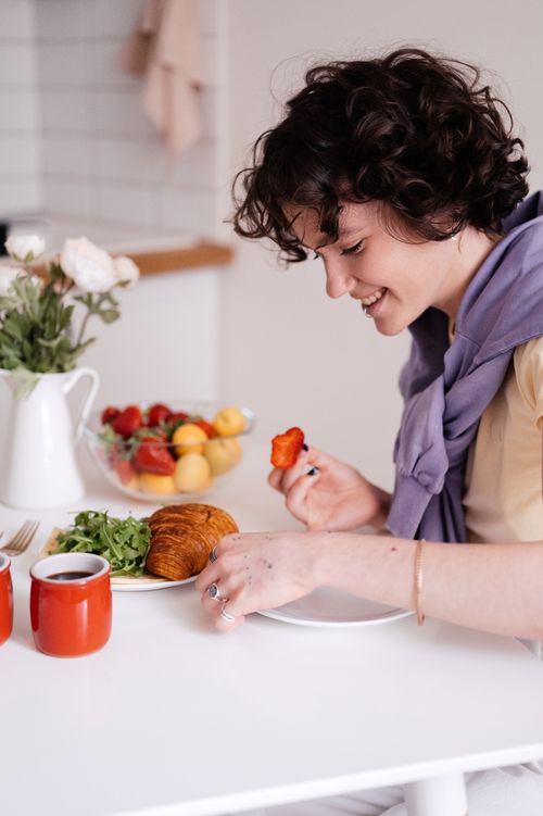 Free A young woman enjoys breakfast with a croissant and coffee in a cozy, naturally lit kitchen setting. Stock Photo