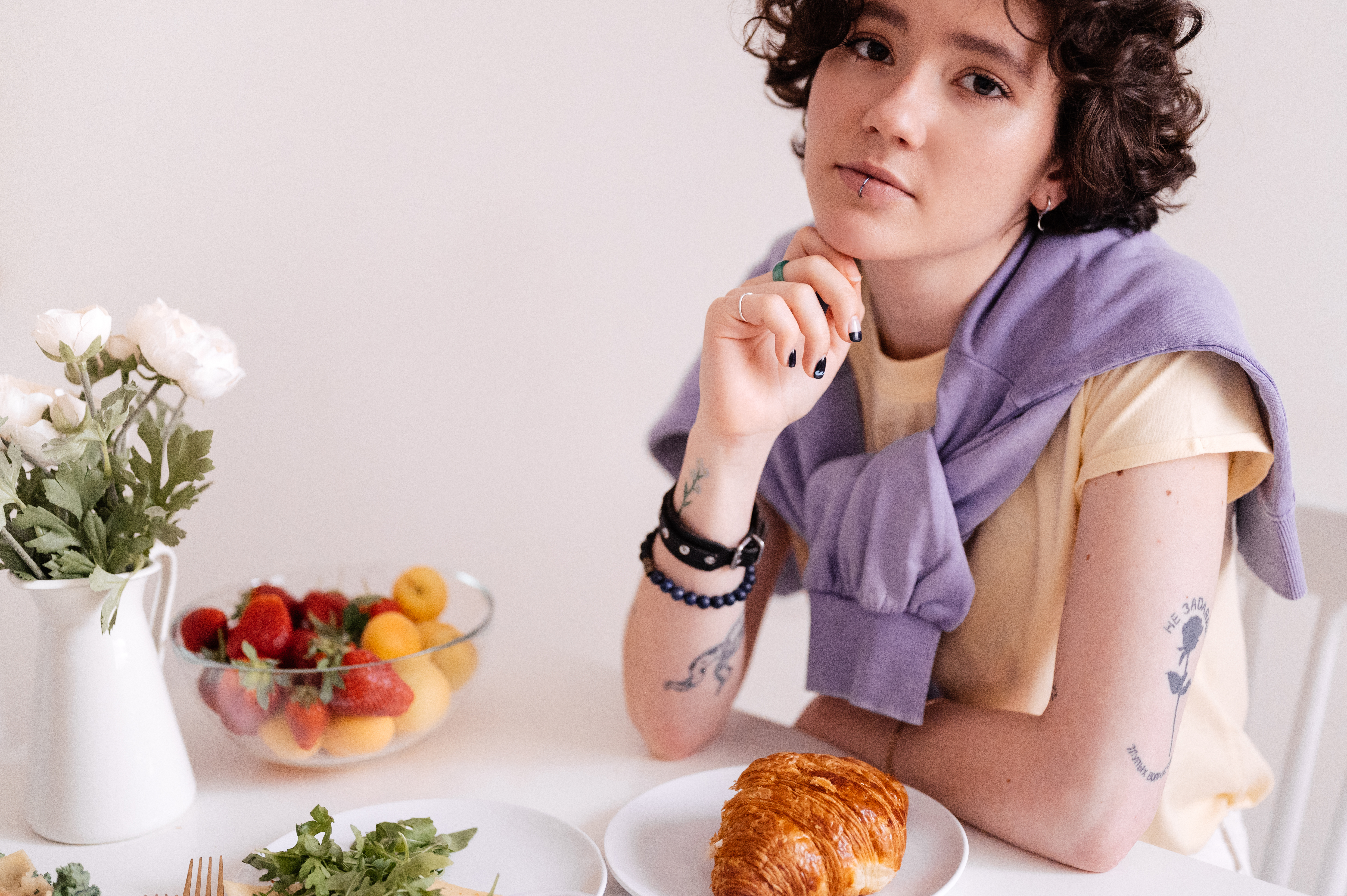 A stylish teenager with piercings enjoys a breakfast of croissant and fruit indoors.