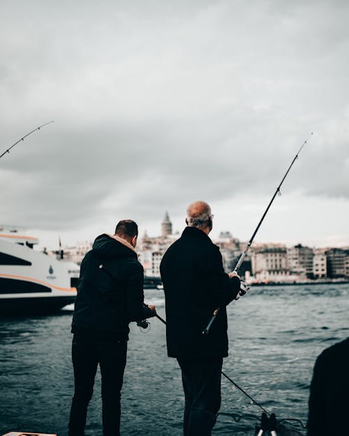 Back view of unrecognizable fisherman in warm clothes standing with fishing rod on embankment near river on cloudy autumn day