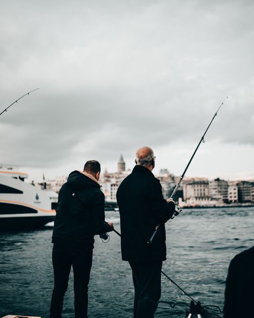 Free Back view of unrecognizable fisherman in warm clothes standing with fishing rod on embankment near river on cloudy autumn day Stock Photo