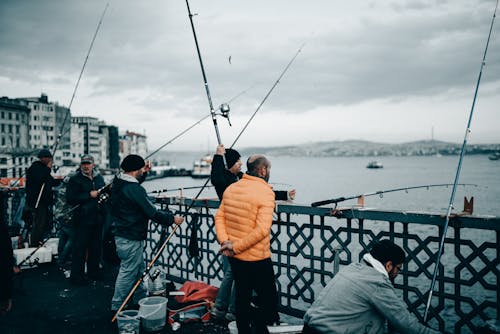 Group of unrecognizable fishermen with fishing rods standing on embankment near sea and buildings under cloudy sky in autumn day
