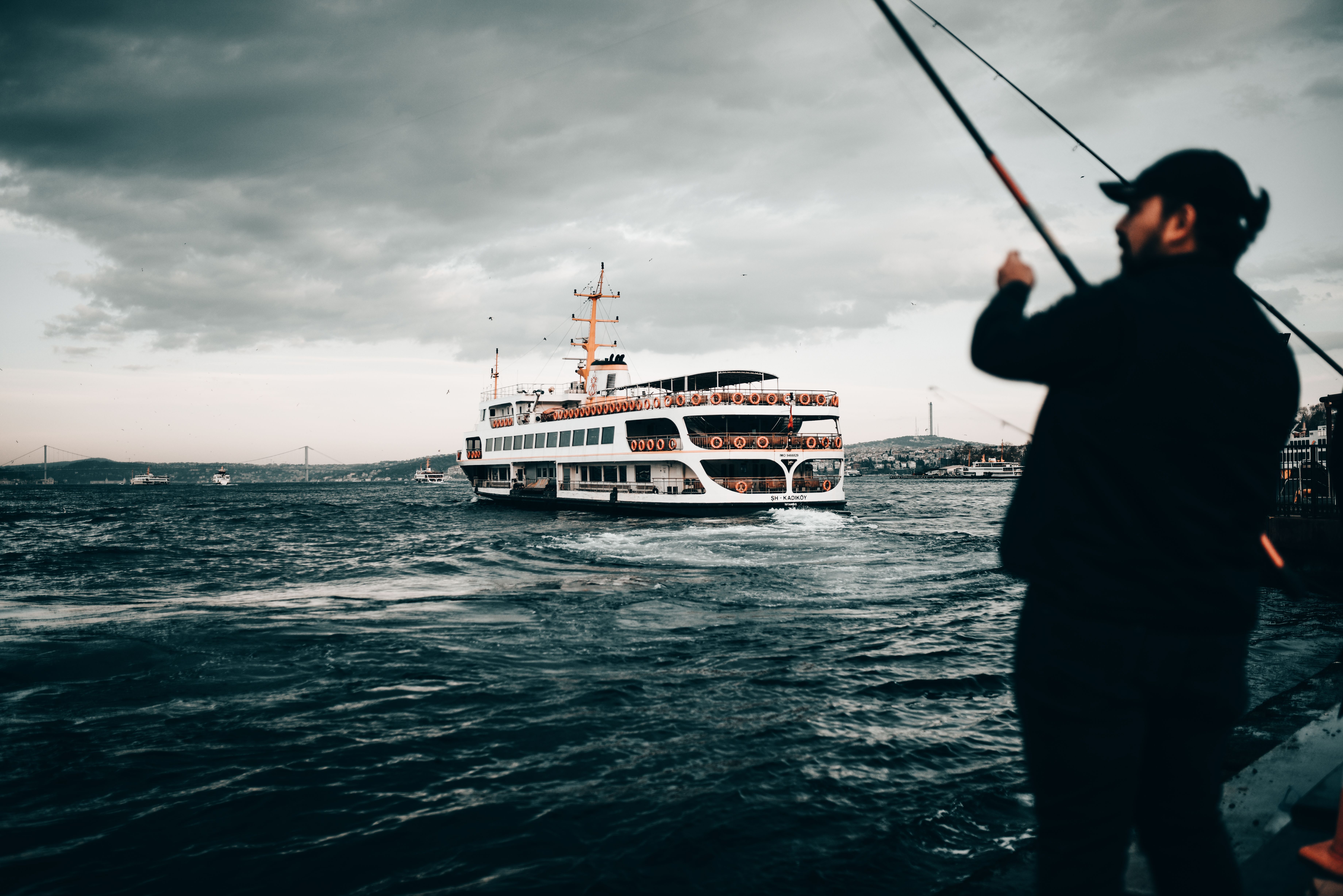 Unrecognizable male with fishing rod standing on embankment of rippling sea with modern yacht and fishing under cloudy sky in evening