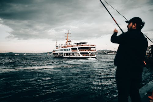 Free Unrecognizable male with fishing rod standing on embankment of rippling sea with modern yacht and fishing under cloudy sky in evening Stock Photo