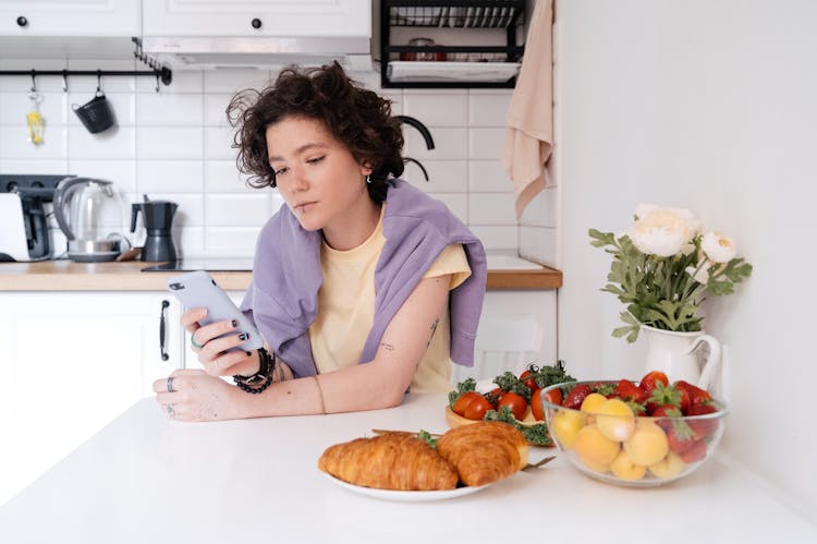 Woman Sitting With Smartphone By Table In Kitchen