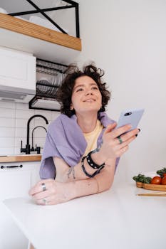 Smiling woman leaning on kitchen counter, holding smartphone, surrounded by fresh vegetables.