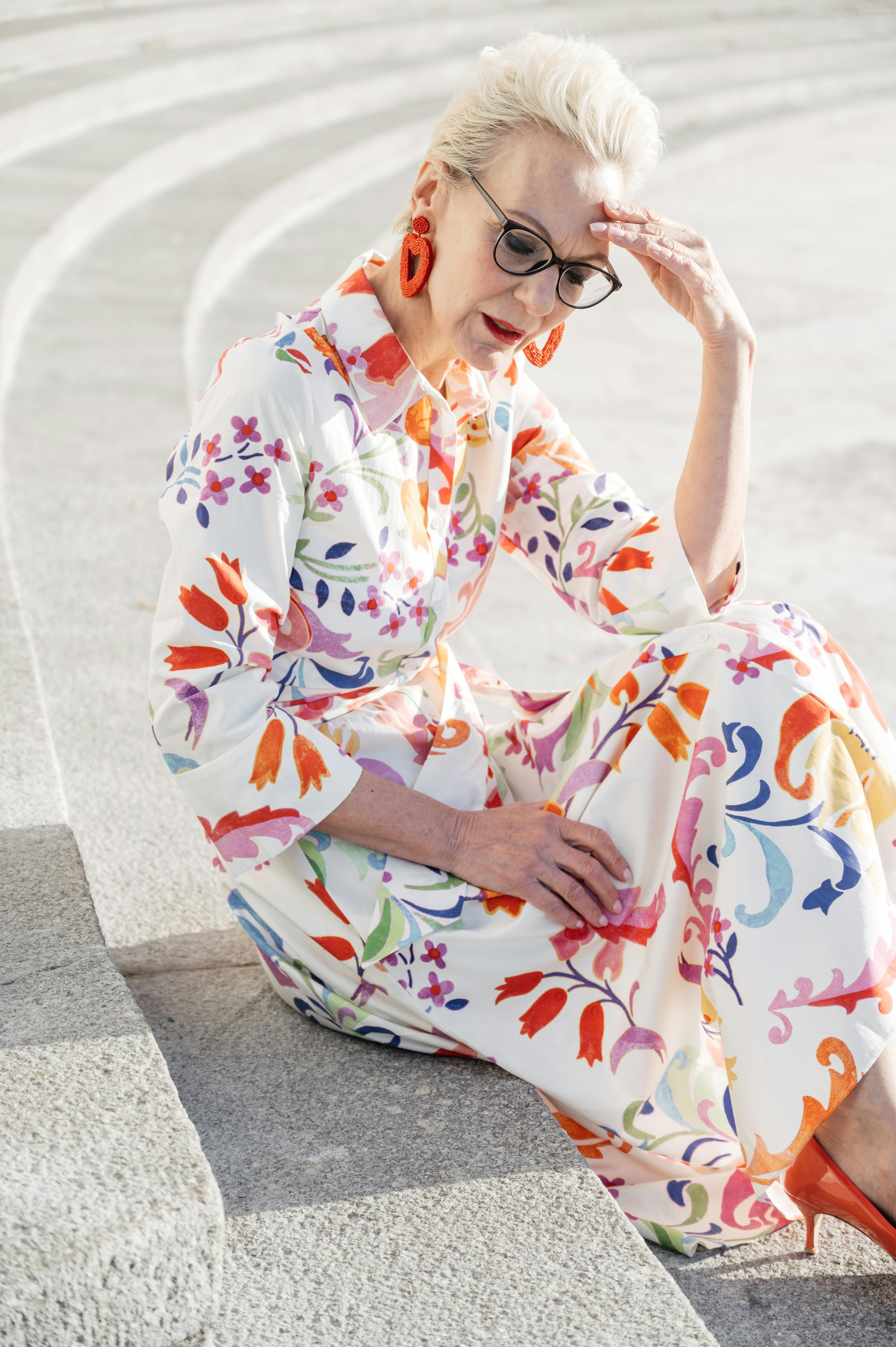 an elderly woman in printed dress sitting on a concrete stairs with her hand on her head