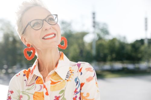 Free Charming senior woman with glasses and heart earrings smiles joyfully outdoors in floral attire. Stock Photo