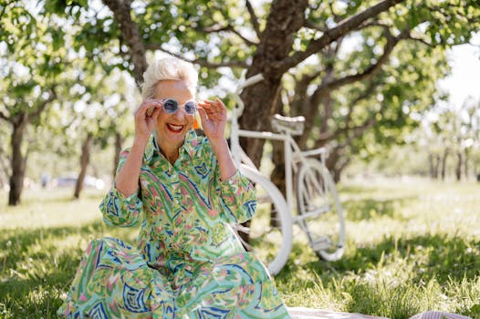 A joyful senior woman in a colorful dress smiles while outdoors, enjoying a sunny day with a bicycle.