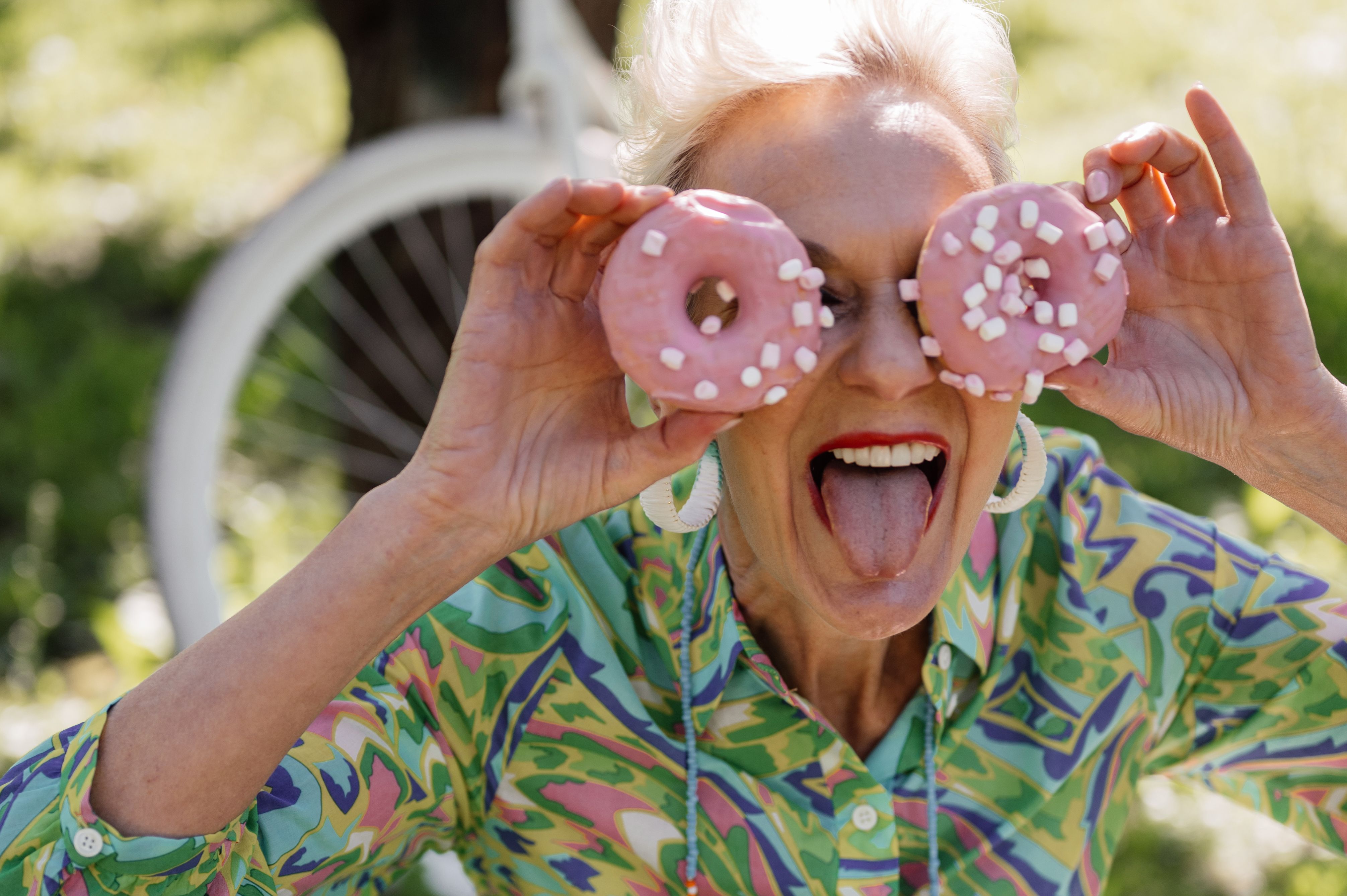 A joyful senior woman outdoors holding pink donuts as glasses, showing playfulness and humor.