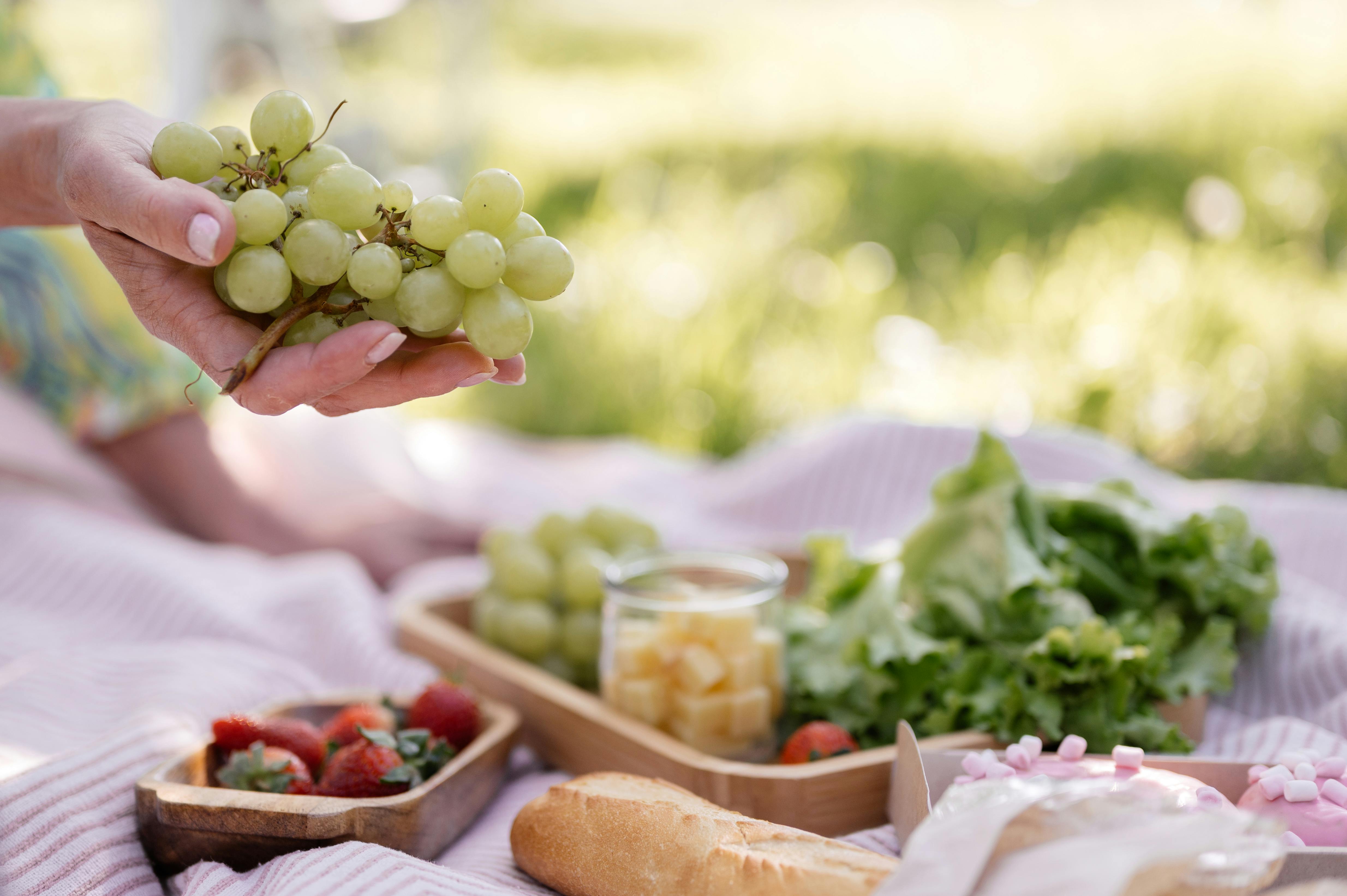 Hand holding fresh grapes at a summer picnic with greens, strawberries, and bread.