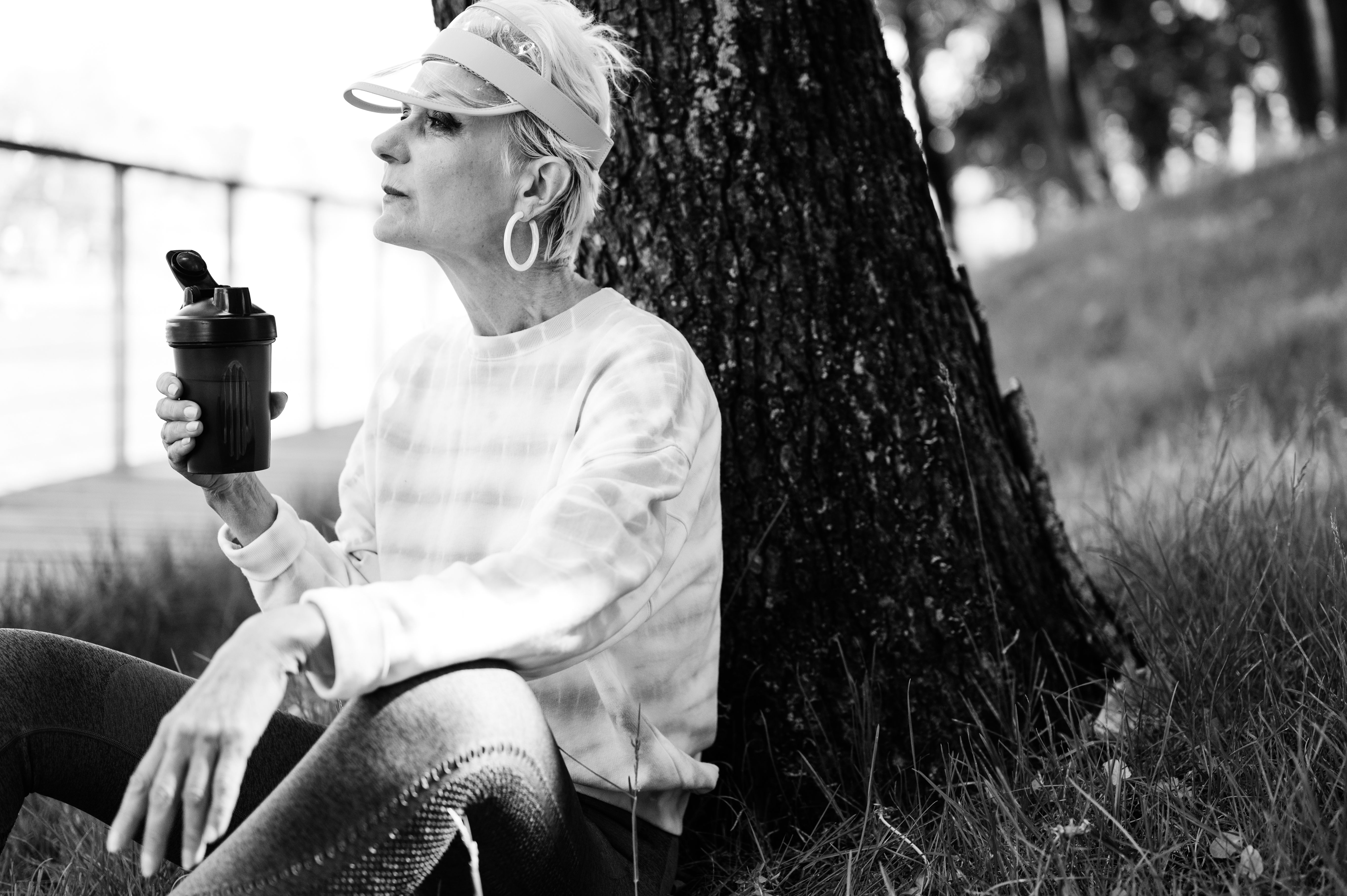 A senior woman relaxes against a tree, enjoying a drink in a park setting.
