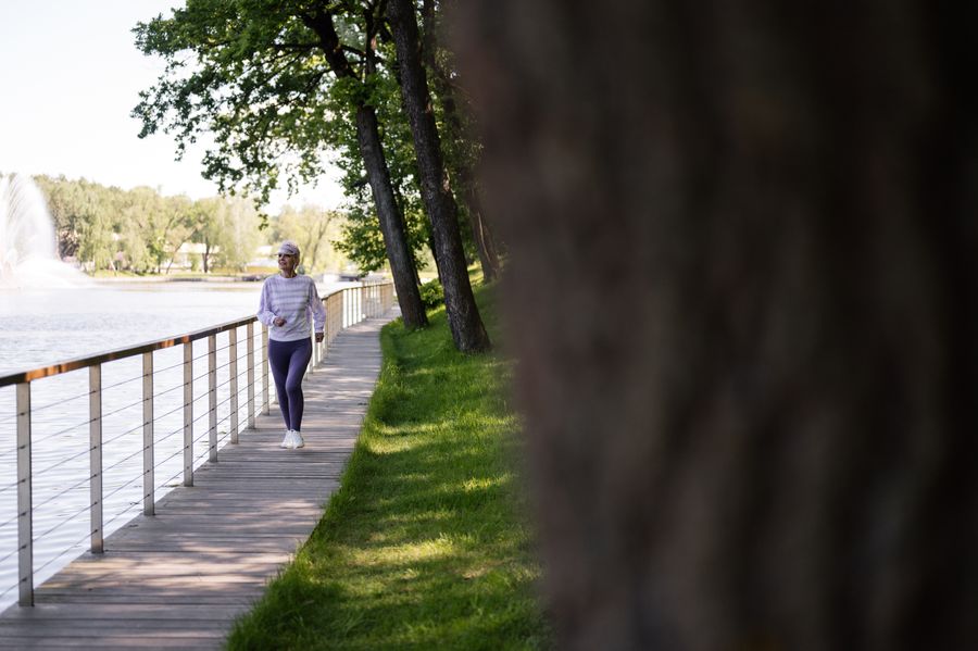 A woman in her fifties walking along a sunny lakeside boardwalk lined with trees.