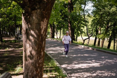 Free A senior woman jogging along a tree-lined path on a sunny day, embracing a healthy lifestyle. Stock Photo
