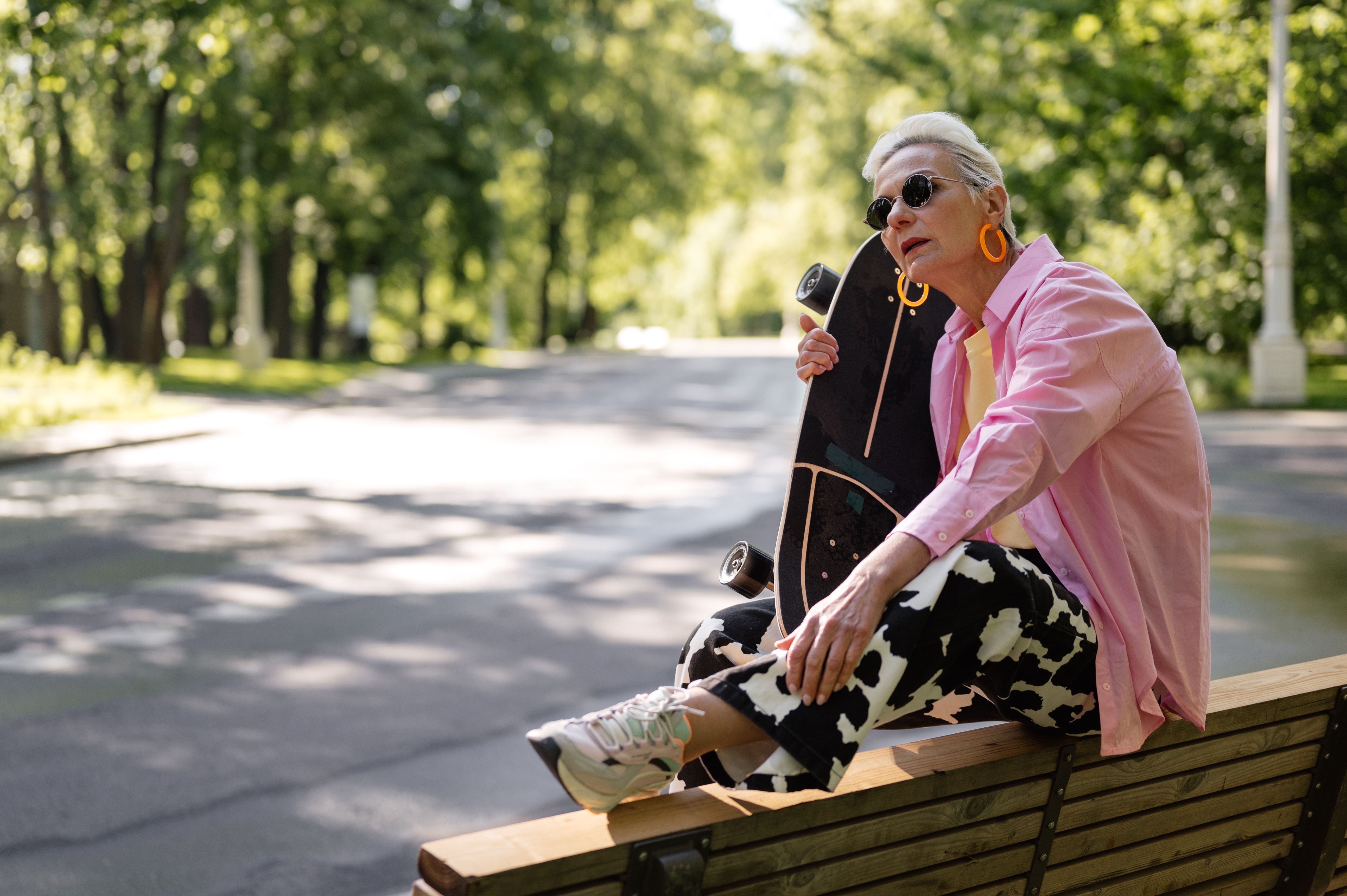 Elderly woman in fashionable attire leaning on skateboard while sitting on a bench in a park.