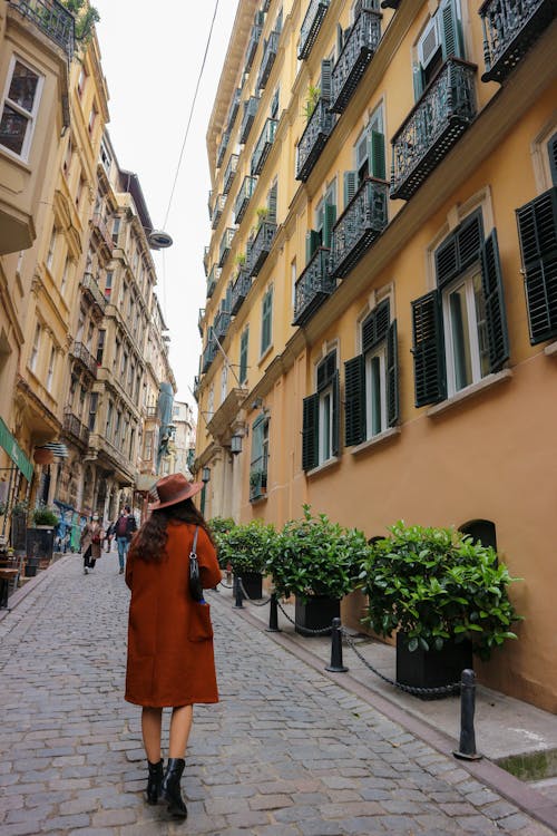Free Woman in Orange Coat Walking at Narrow City Street Stock Photo