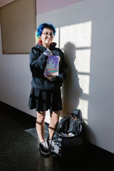Young student with vibrant hair holding a book in a well-lit school hallway, embracing a unique style.