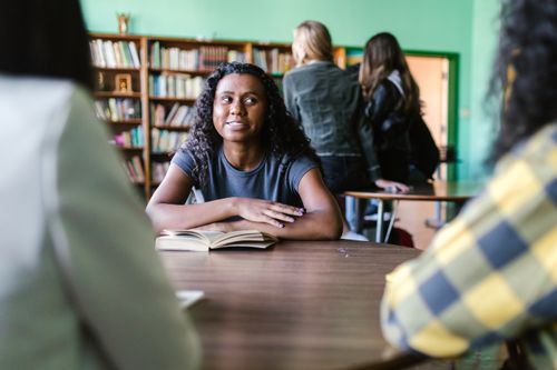 Free Young woman with curly hair studying in a cozy library environment, surrounded by books and peers. Stock Photo