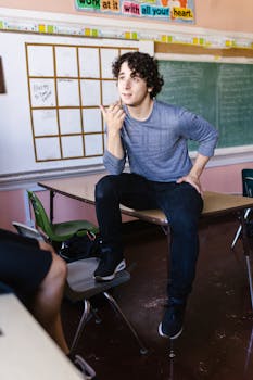 A young man with curly hair sits on a classroom desk, talking animatedly to a fellow student.