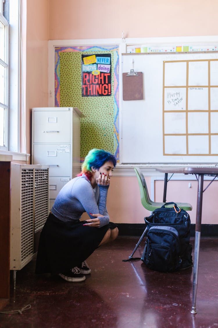 A Woman With Colorful Hair Sitting On The Floor Of A Classroom While In Deep Thought