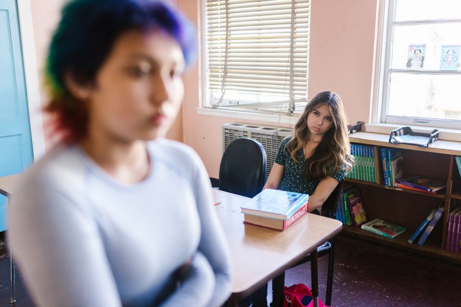 Two teenage girls in a classroom setting, one focused, the other blurred in foreground.