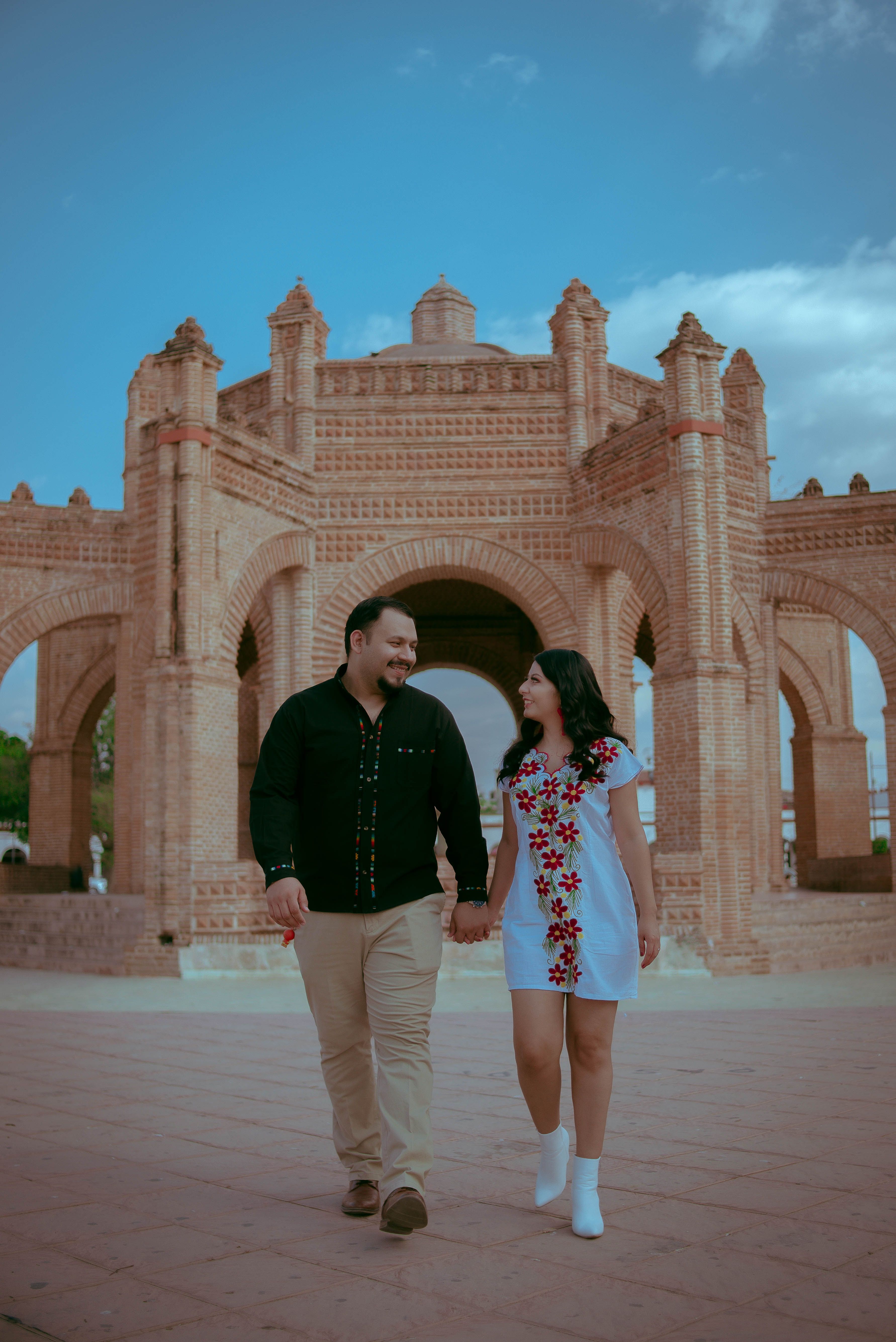 A couple strolls hand in hand near a historic brick landmark on a clear day.
