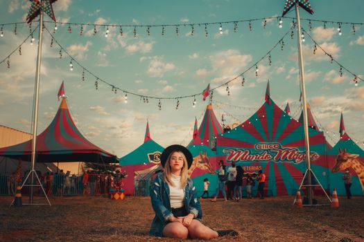 A young woman with a hat sits in front of a colorful vintage circus tent under string lights during sunset.
