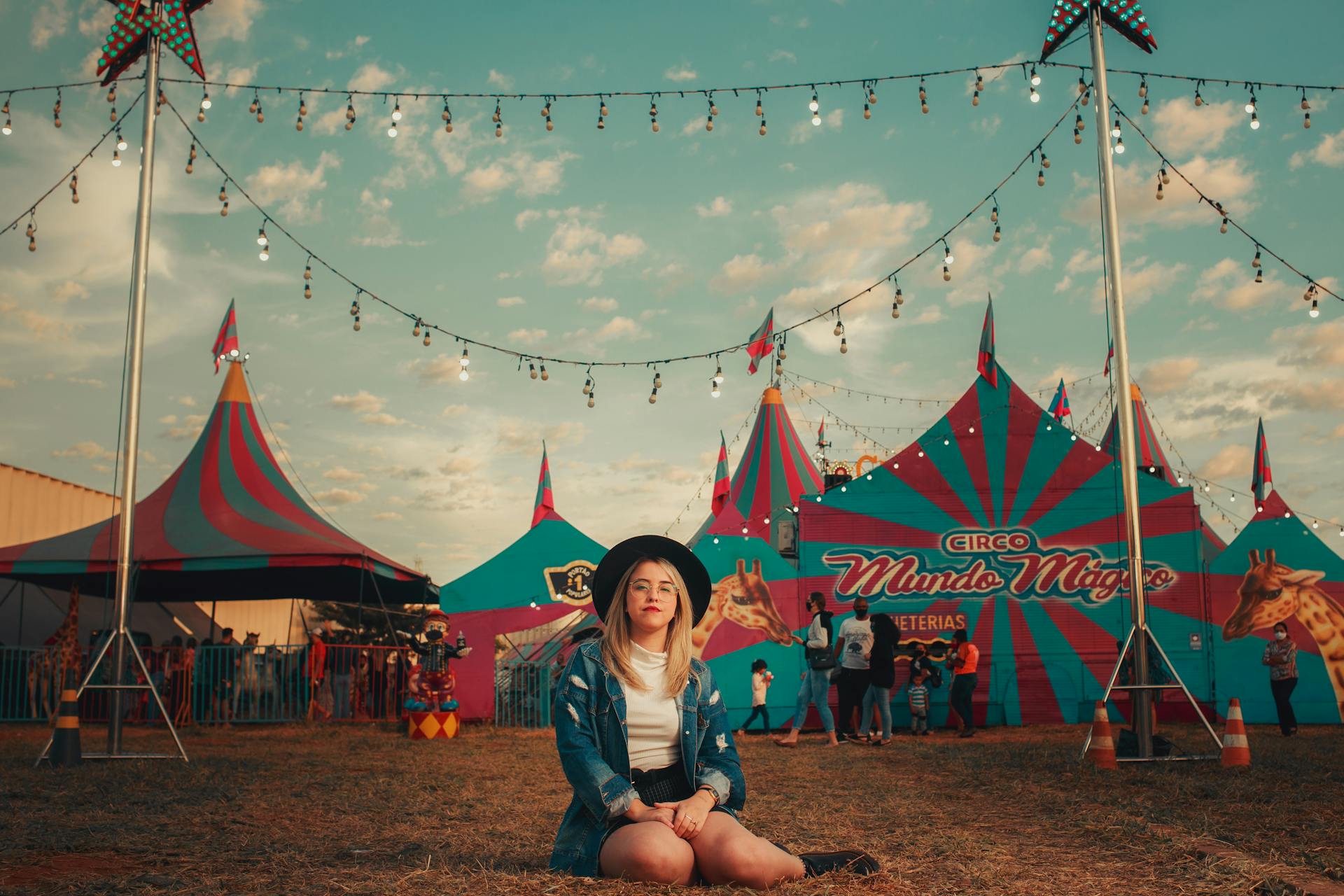 A young woman with a hat sits in front of a colorful vintage circus tent under string lights during sunset.