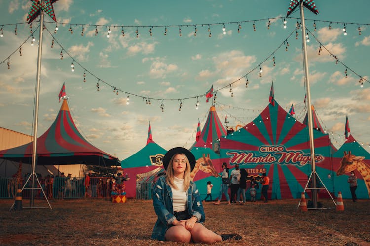 Woman In Hat Sitting On Grass Near Circus