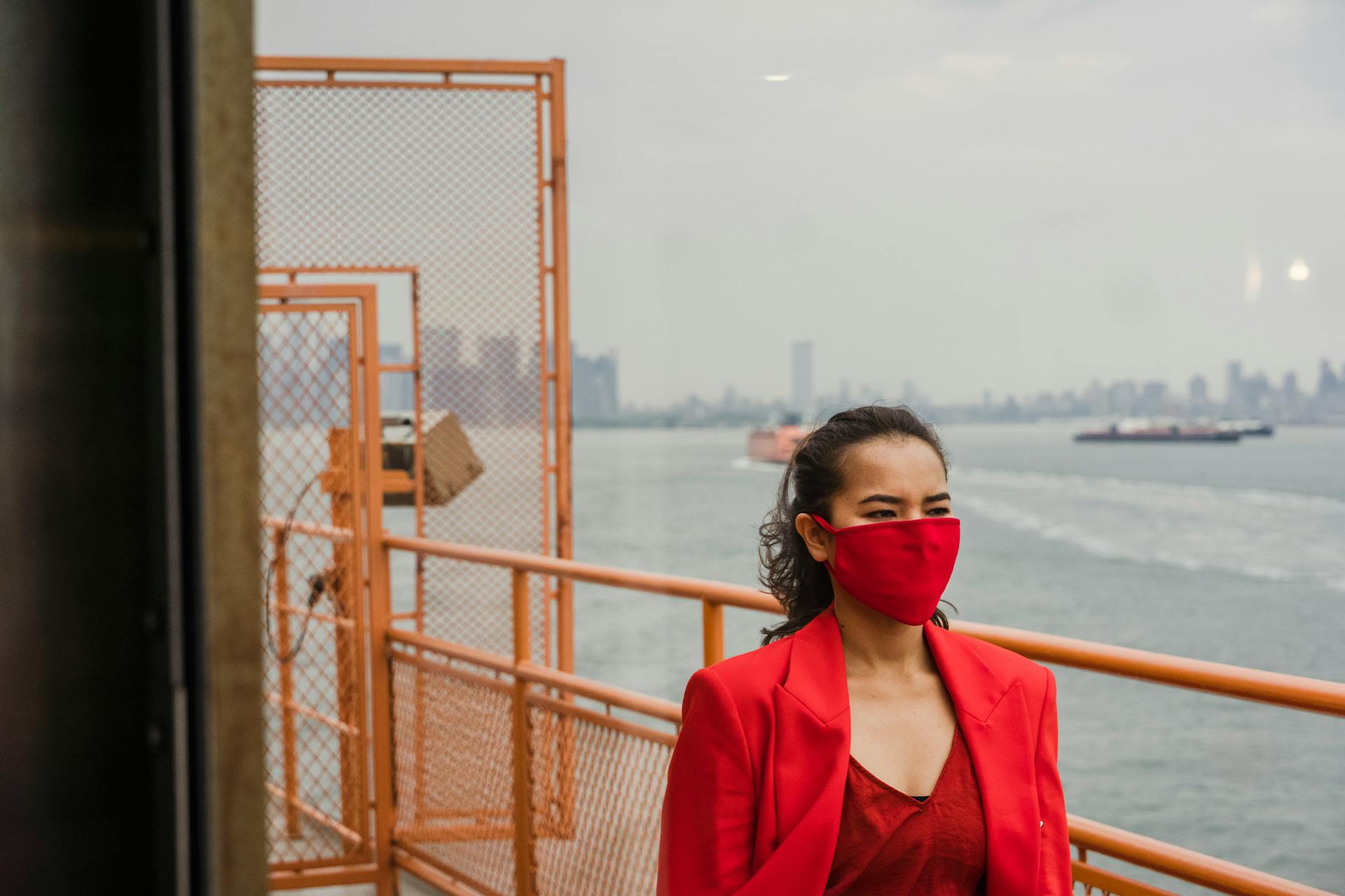 A woman in a red outfit and mask stands on a ferry with a city skyline in the background.