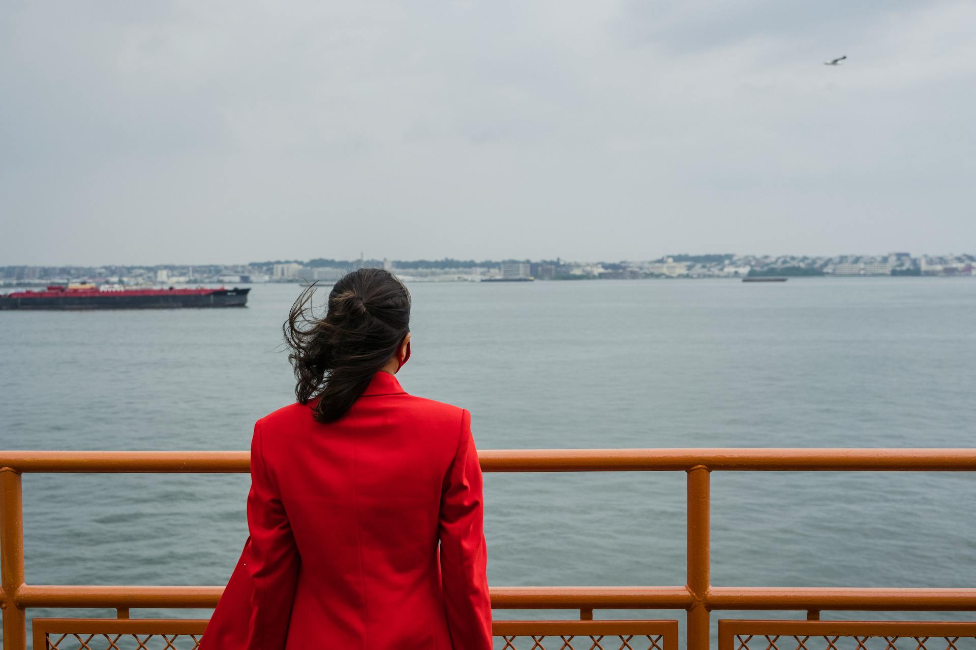Woman in a red coat looking at the ocean from a ferry observation deck.