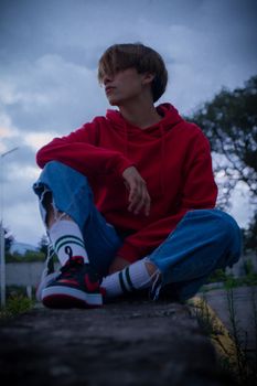 Young person in casual attire sitting on a stone wall in an outdoor setting under a cloudy sky.