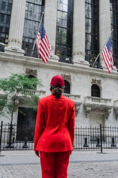 A woman in red stands facing a historic Wall Street building with American flags.