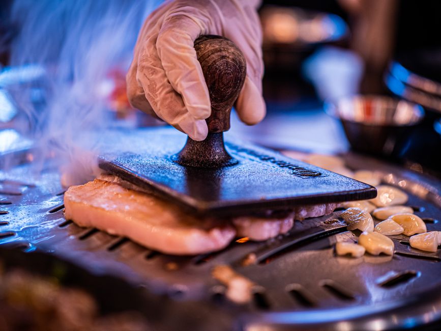 A close-up image of a gloved hand pressing meat on a grill with fresh garlic, capturing a tasty cooking moment.