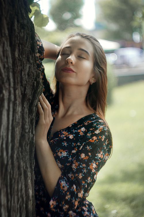 Free Woman in floral dress enjoying a peaceful moment with closed eyes by a tree in a sunny park. Stock Photo