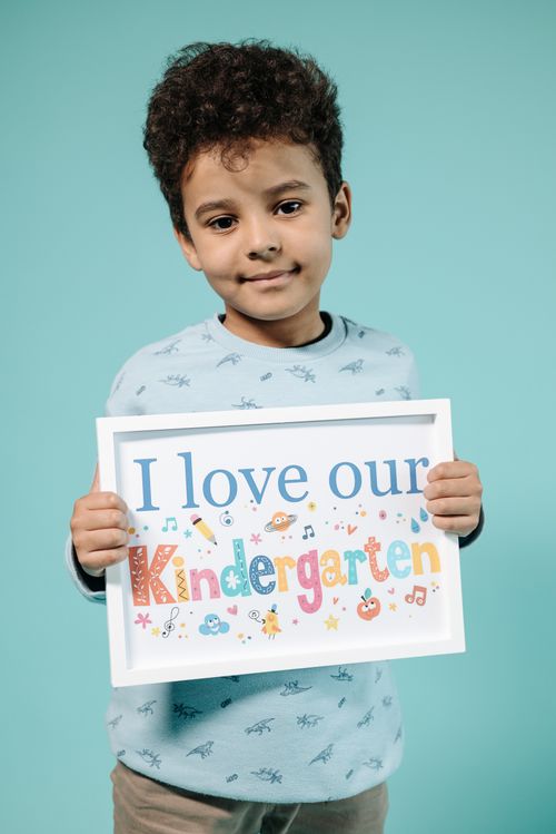 Free Cheerful child holding a colorful kindergarten sign against a blue backdrop. Stock Photo