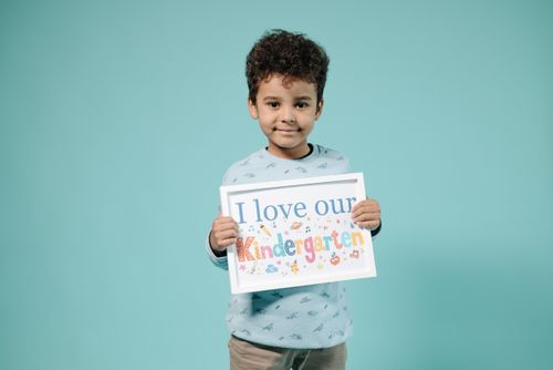 Free Young child with curly hair holding a sign that says 'I love our Kindergarten' against a turquoise background. Stock Photo