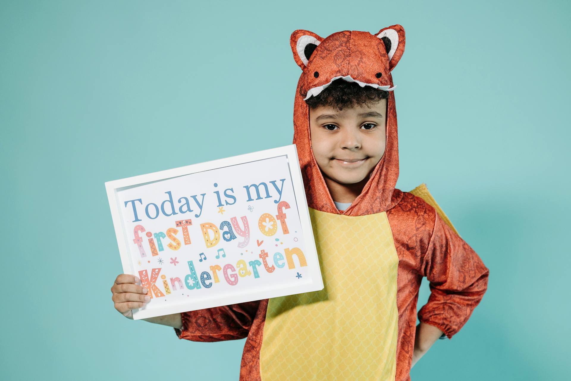 Smiling child in a dinosaur costume holding a first day of kindergarten sign against a pastel background.