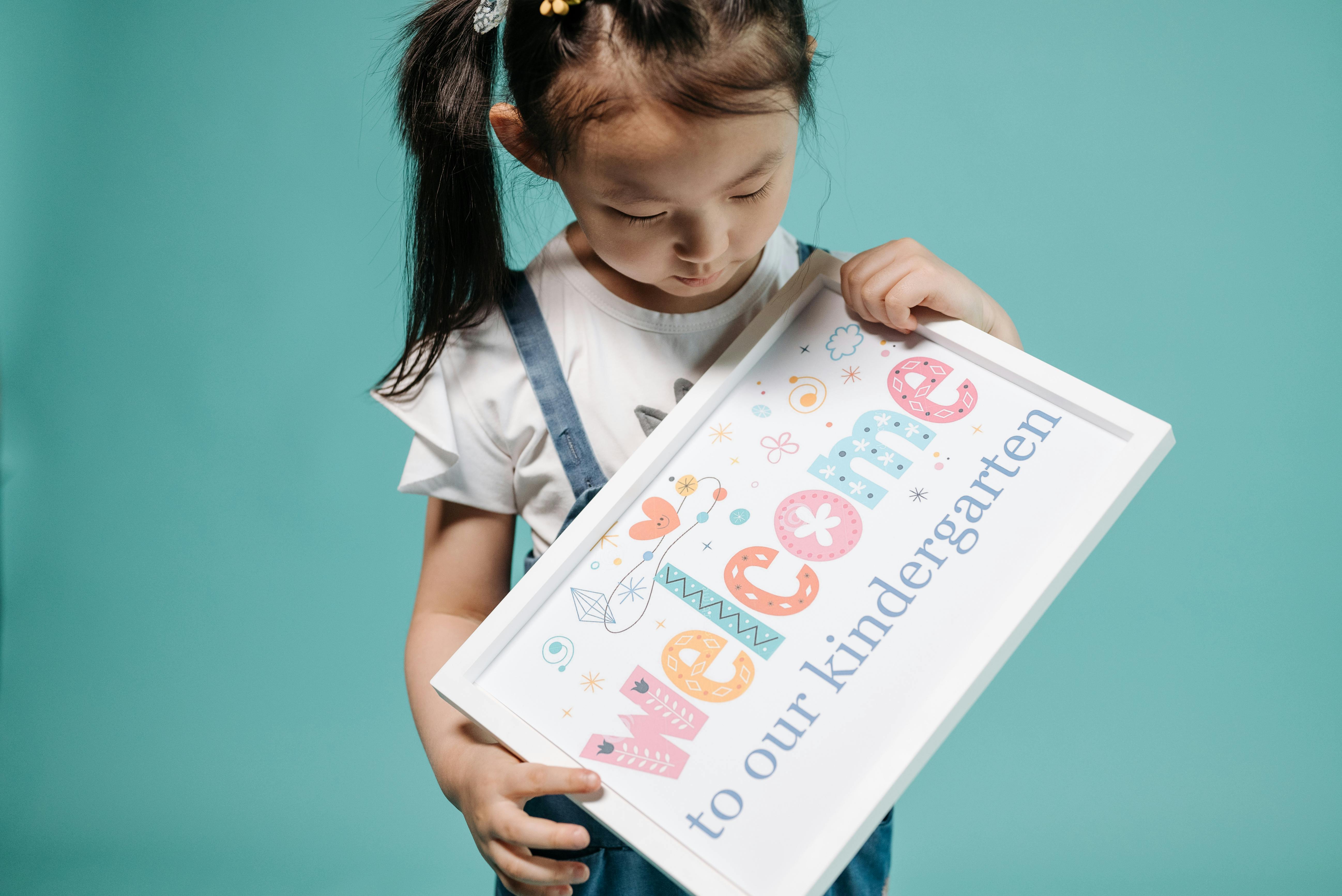 A young girl in overalls holds a colorful kindergarten welcome sign.