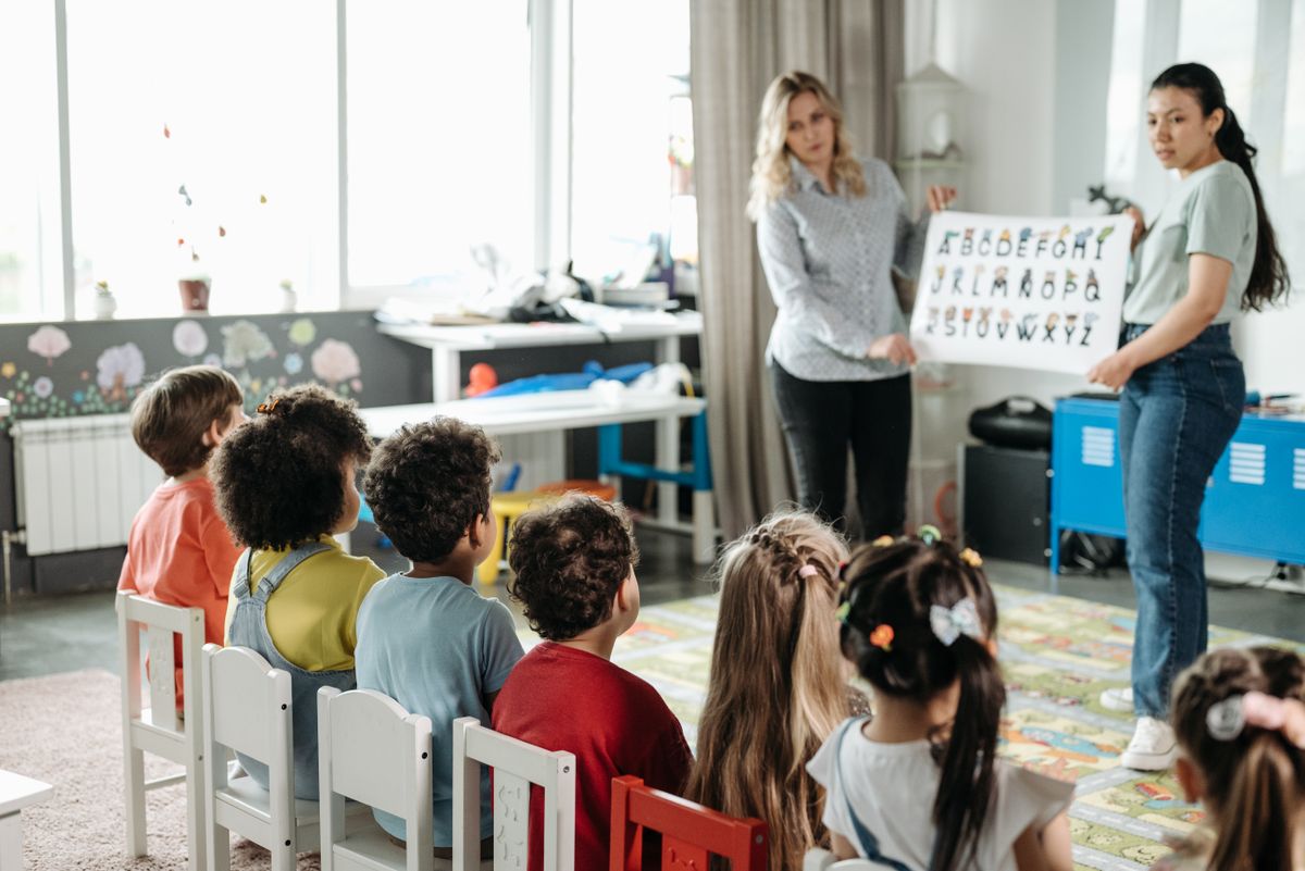Children seated in a row facing their teachers at an alphabet board.