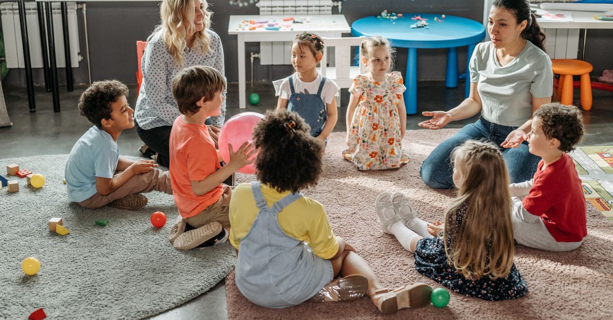 A diverse group of children sitting in a circle, listening to a teacher with colorful educational materials in the background