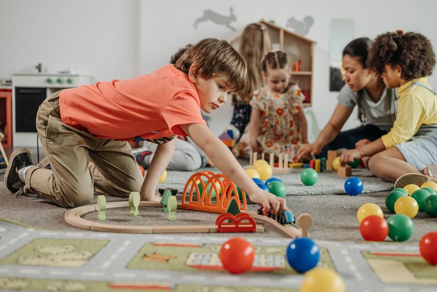 A boy guiding a wooden train along a track while a teacher plays nearby