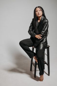 Fashionable woman with braided hair in a leather jacket sitting on a stool, exuding confidence in an indoor studio setting.