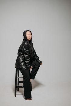 Asian woman with braids poses barefoot on stool in a studio setting wearing black leather jacket.