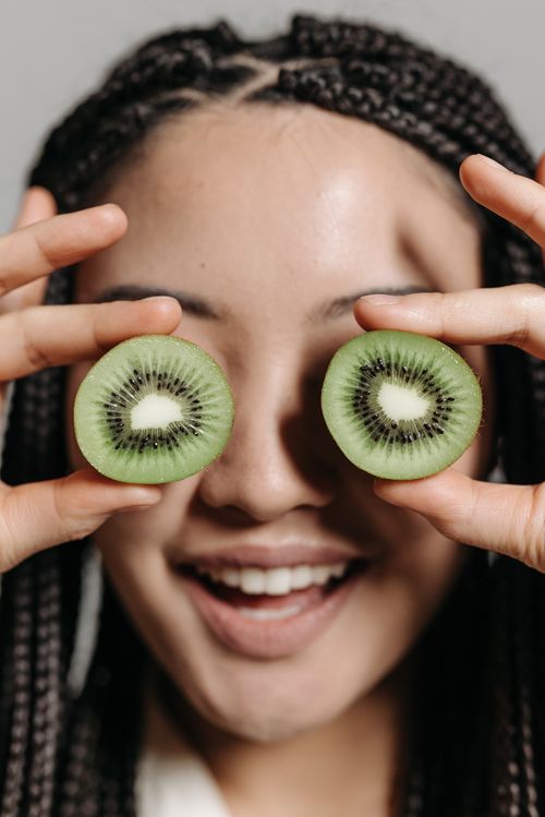 Free Young woman smiling while playfully holding kiwi slices in front of her eyes. Stock Photo